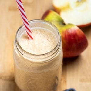 An apple smoothie in a mason jar with a straw sitting on a wooden cutting board with a teaspoon of cinnamon in front of it, and an apple cut in half behind it.