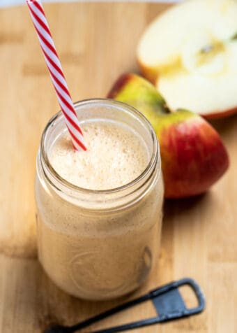 An apple smoothie in a mason jar with a straw sitting on a wooden cutting board with a teaspoon of cinnamon in front of it, and an apple cut in half behind it.