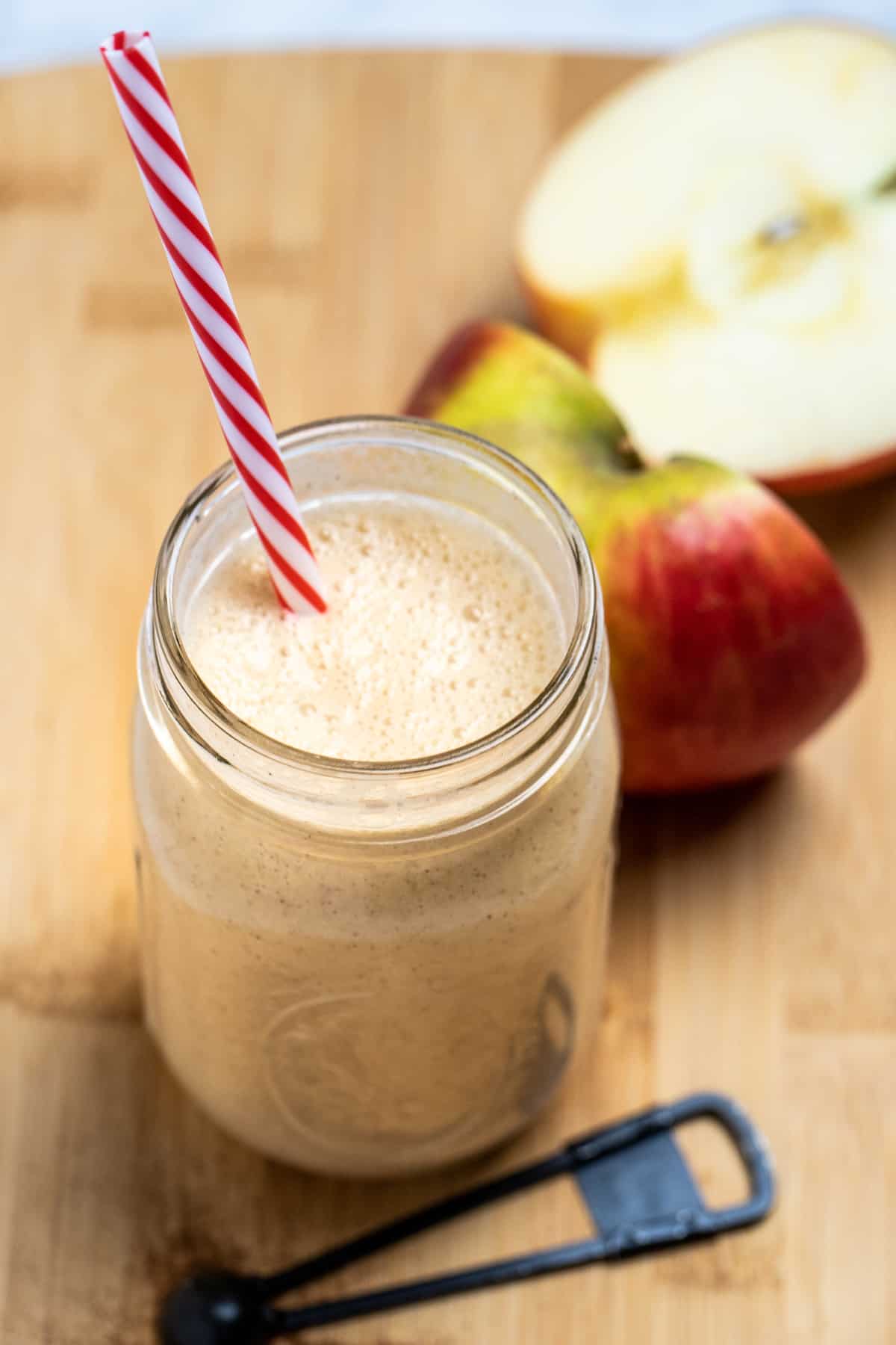 An apple smoothie in a mason jar with a straw sitting on a wooden cutting board with a teaspoon of cinnamon in front of it, and an apple cut in half behind it.