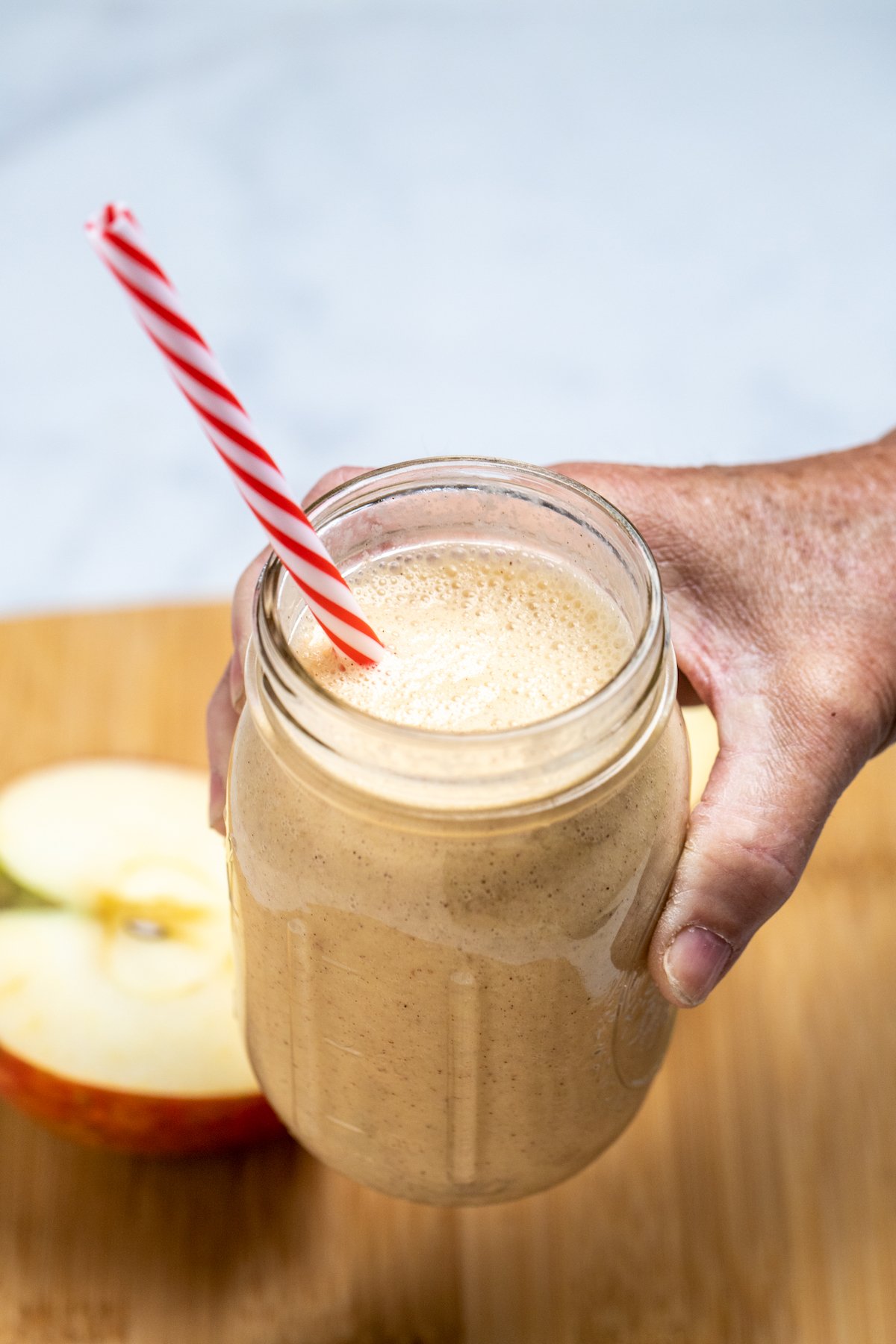A hand holding an apple banana smoothie in a mason jar with a straw above a wooden cutting board with an apple cut in half behind it.
