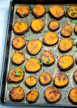 Sweet potato slices on a sheet pan with parchment paper, fully cooked with caramelized tops and fresh parsley on top.