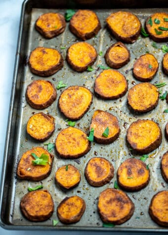 Sweet potato slices on a sheet pan with parchment paper, fully cooked with caramelized tops and fresh parsley on top.