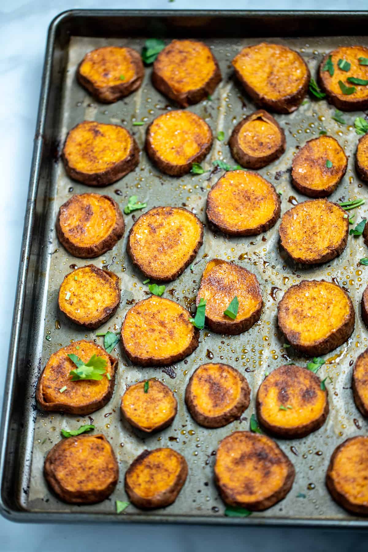 Sweet potato slices on a sheet pan with parchment paper, fully cooked with caramelized tops and fresh parsley on top.