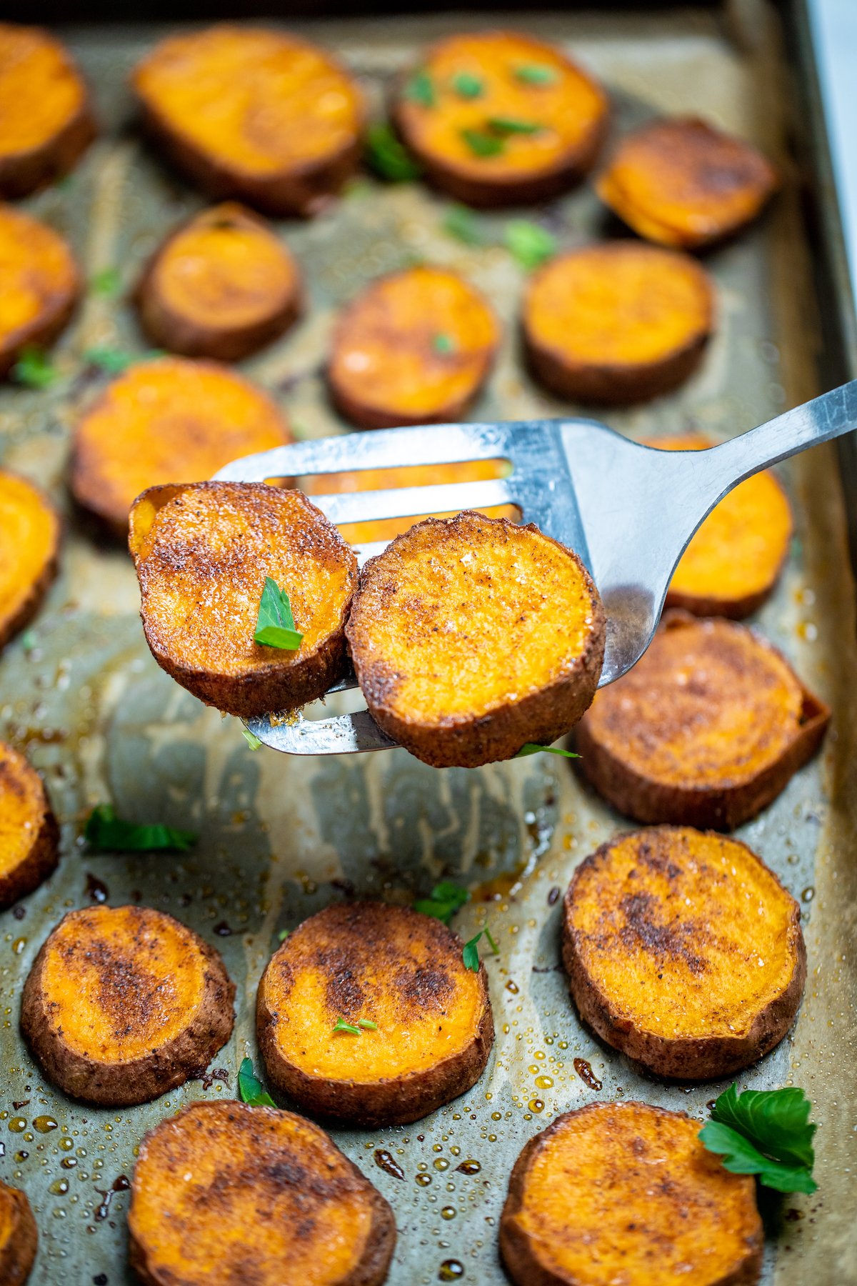 A spatula lifting up two sweet potato slices, above a parchment lined sheet pan with caramelized sweet potato rounds and fresh parsley.
