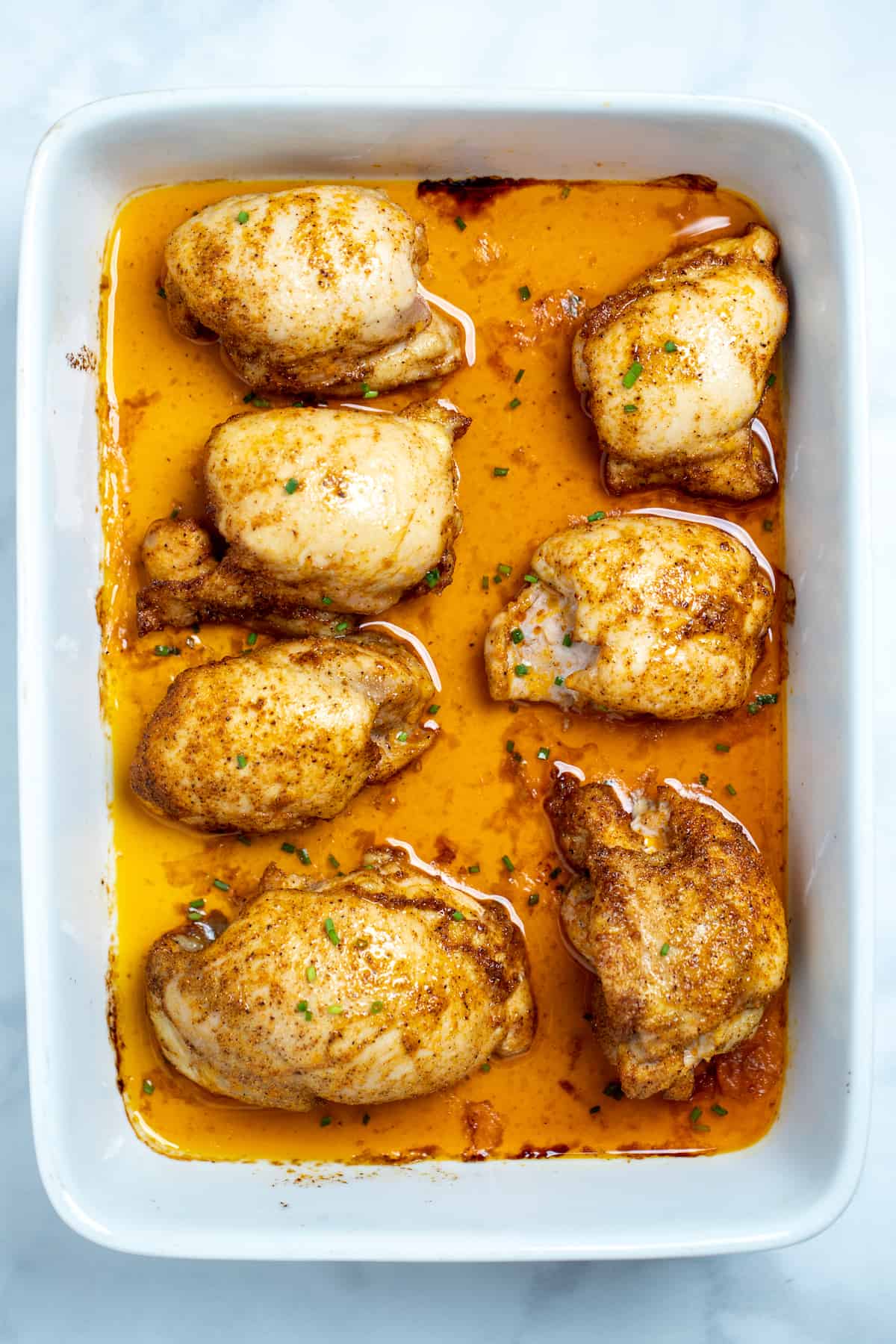 A baking dish of fully cooked chicken thighs sitting in chicken broth, topped with seasoning and fresh chives.