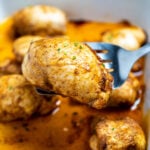 A baking dish of fully cooked chicken thighs sitting in chicken broth, with a spatula lifting one up, topped with seasoning and fresh chives.