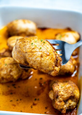 A baking dish of fully cooked chicken thighs sitting in chicken broth, with a spatula lifting one up, topped with seasoning and fresh chives.