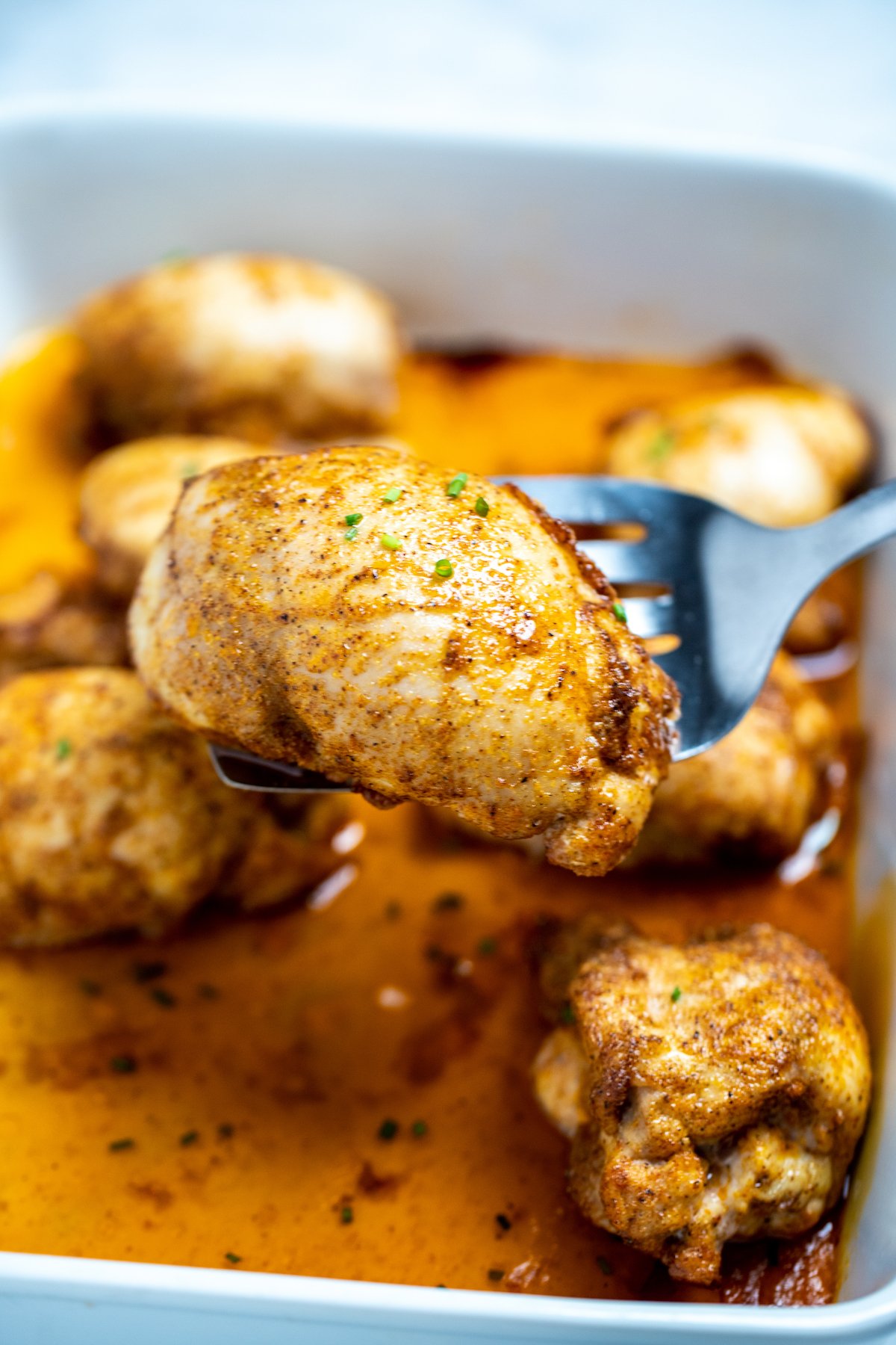 A baking dish of fully cooked chicken thighs sitting in chicken broth, with a spatula lifting one up, topped with seasoning and fresh chives.