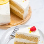 A lemon cake topped with lemon slices, sprinkles, and raspberries on a wooden cake stand, with a slice in front of the cake on a plate with a fork.