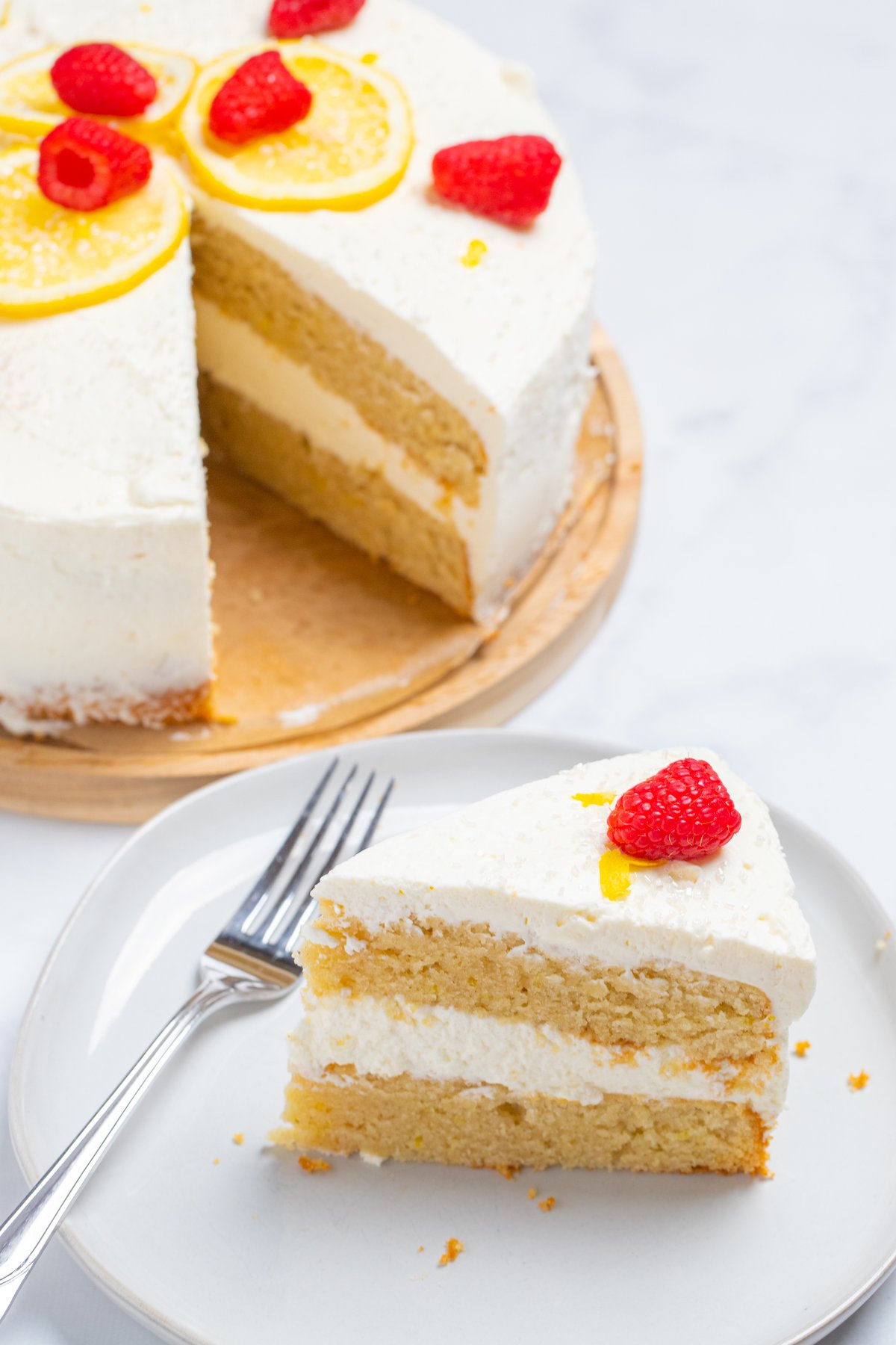 A lemon cake topped with lemon slices, sprinkles, and raspberries on a wooden cake stand, with a slice in front of the cake on a plate with a fork.