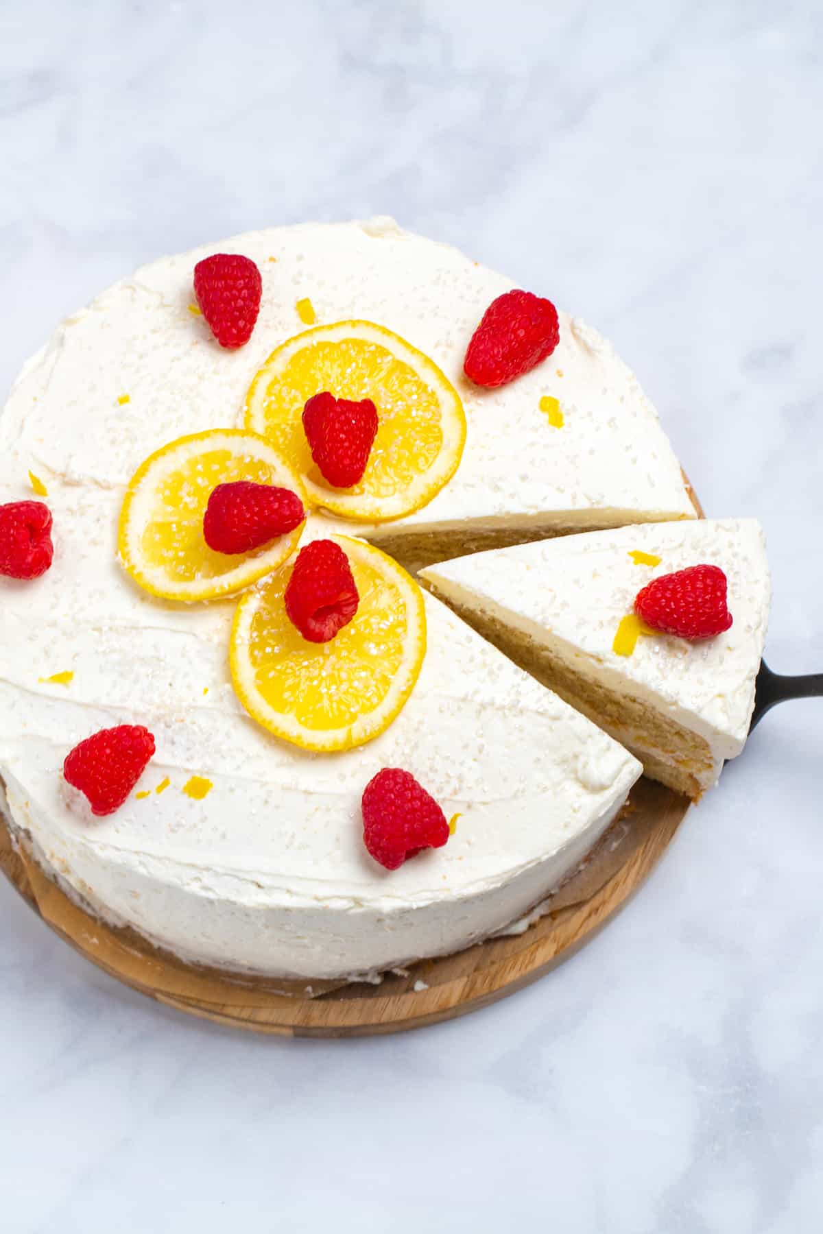 A lemon cake topped with lemon slices, sprinkles, and raspberries on a wooden cake stand, with a slice being lifted out of it.