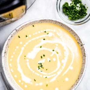 A bowl of cauliflower soup on a table in front of an instant pot, with cream drizzled on top and fresh chives.
