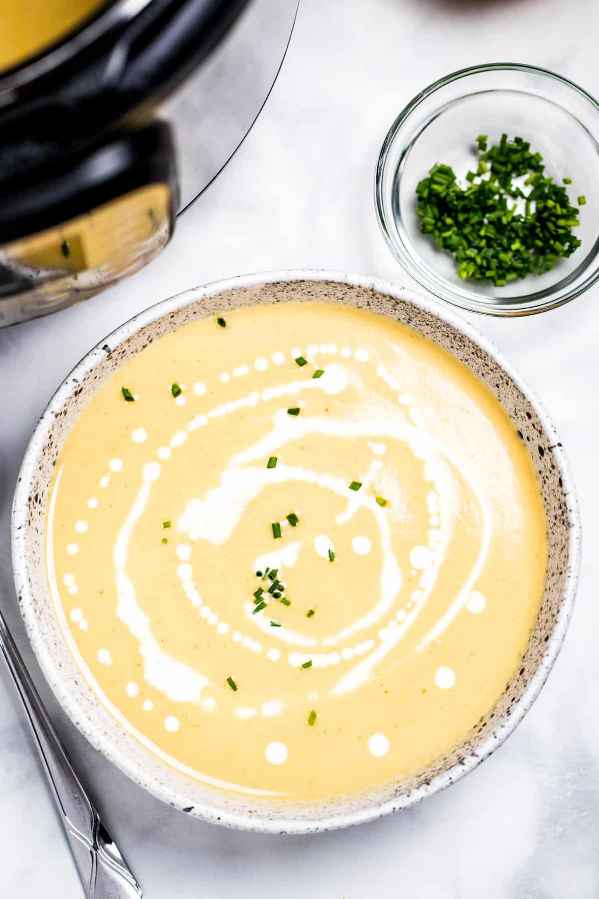 A bowl of cauliflower soup on a table in front of an instant pot, with cream drizzled on top and fresh chives.