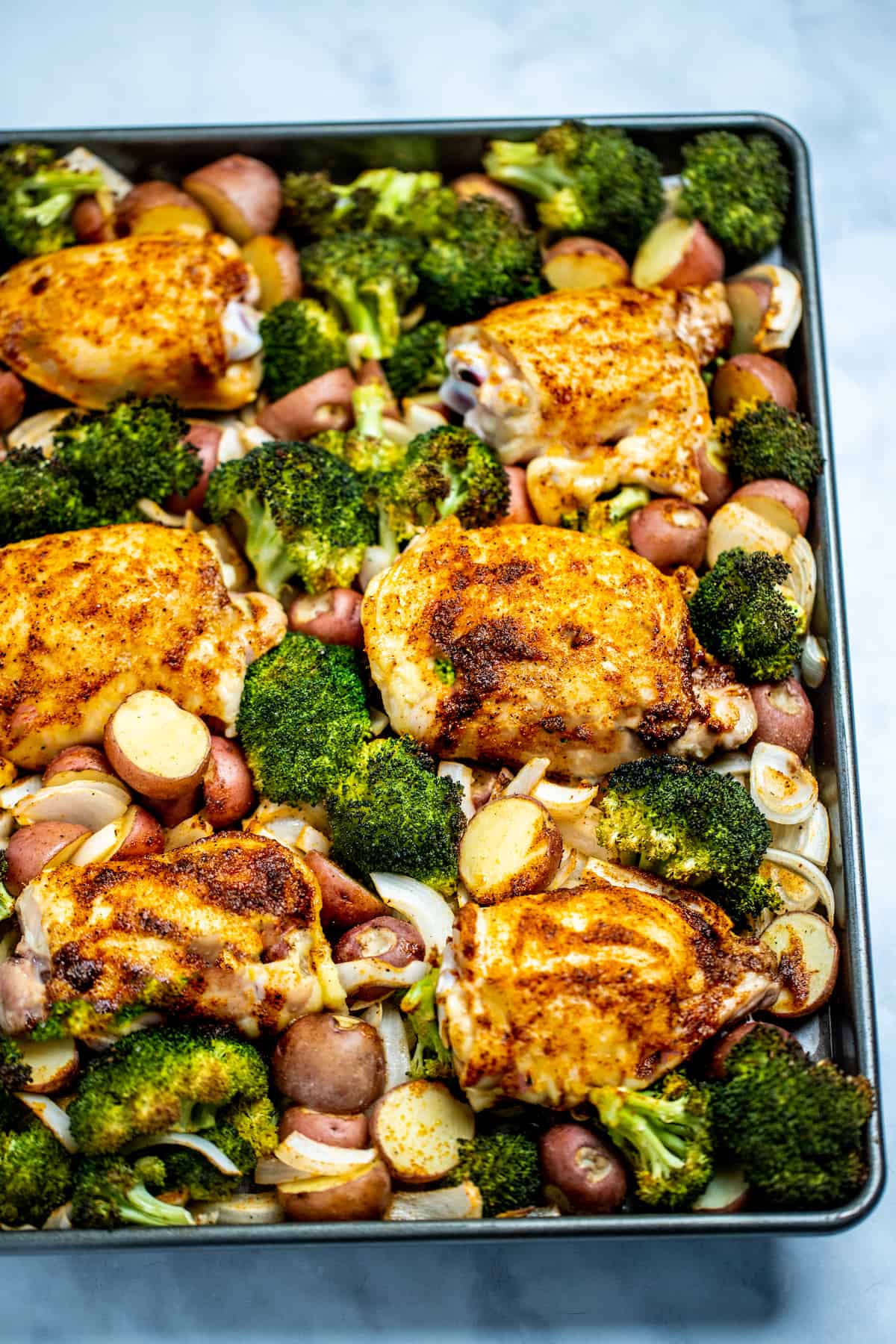 A sheet pan with broccoli, potatoes, sliced onions, and chicken thighs fully cooked and golden brown.