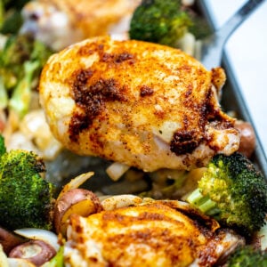 A sheet pan of broccoli, potatoes, onion, and chicken thighs with golden brown skin and a spatula lifting one up.