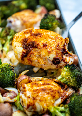 A sheet pan of broccoli, potatoes, onion, and chicken thighs with golden brown skin and a spatula lifting one up.