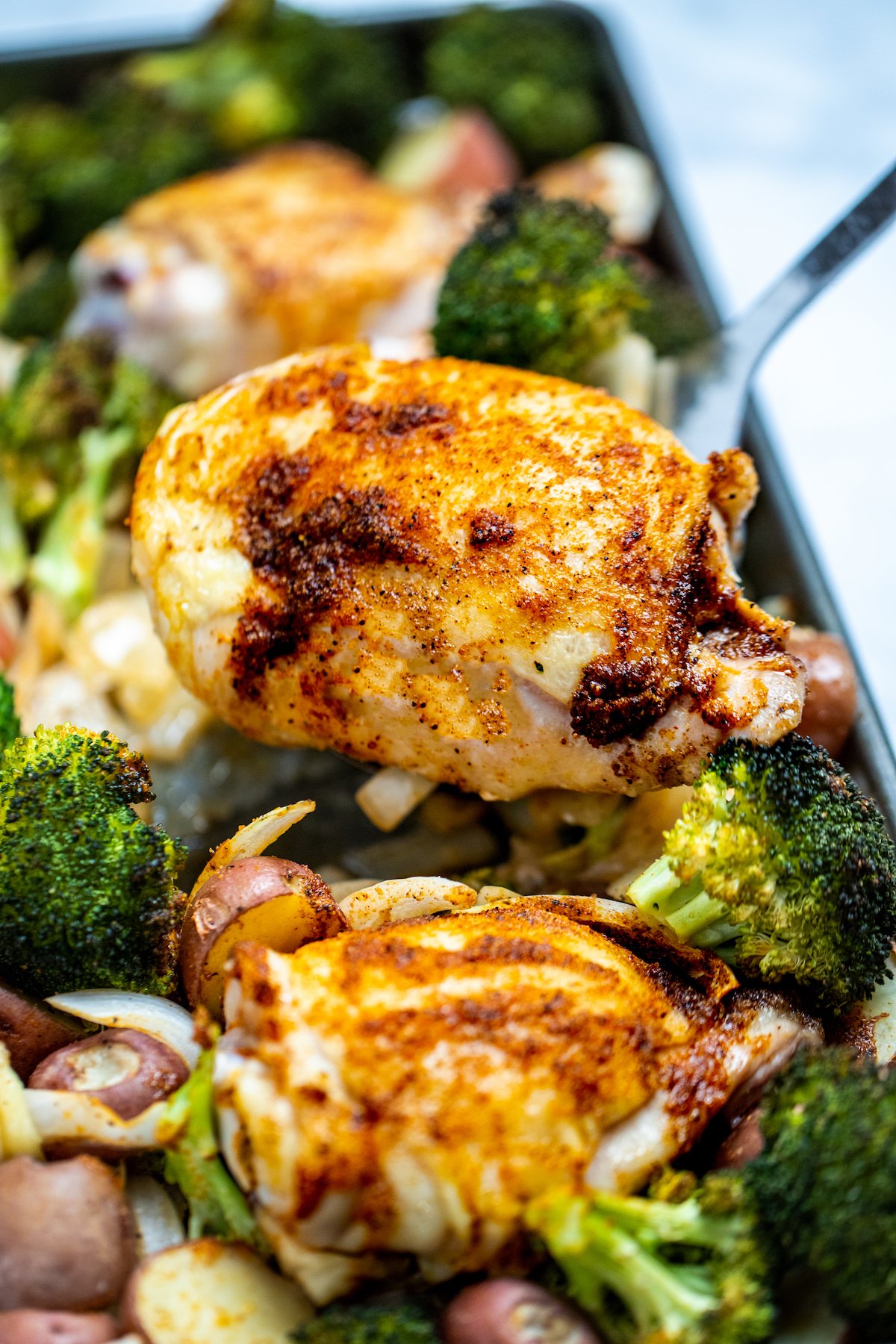 A sheet pan of broccoli, potatoes, onion, and chicken thighs with golden brown skin and a spatula lifting one up.