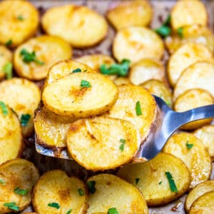 A spatula lifting baked potato slices from a sheet pan, topped with fresh parsley.