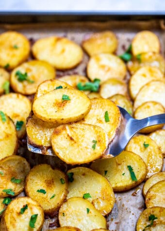 A spatula lifting baked potato slices from a sheet pan, topped with fresh parsley.
