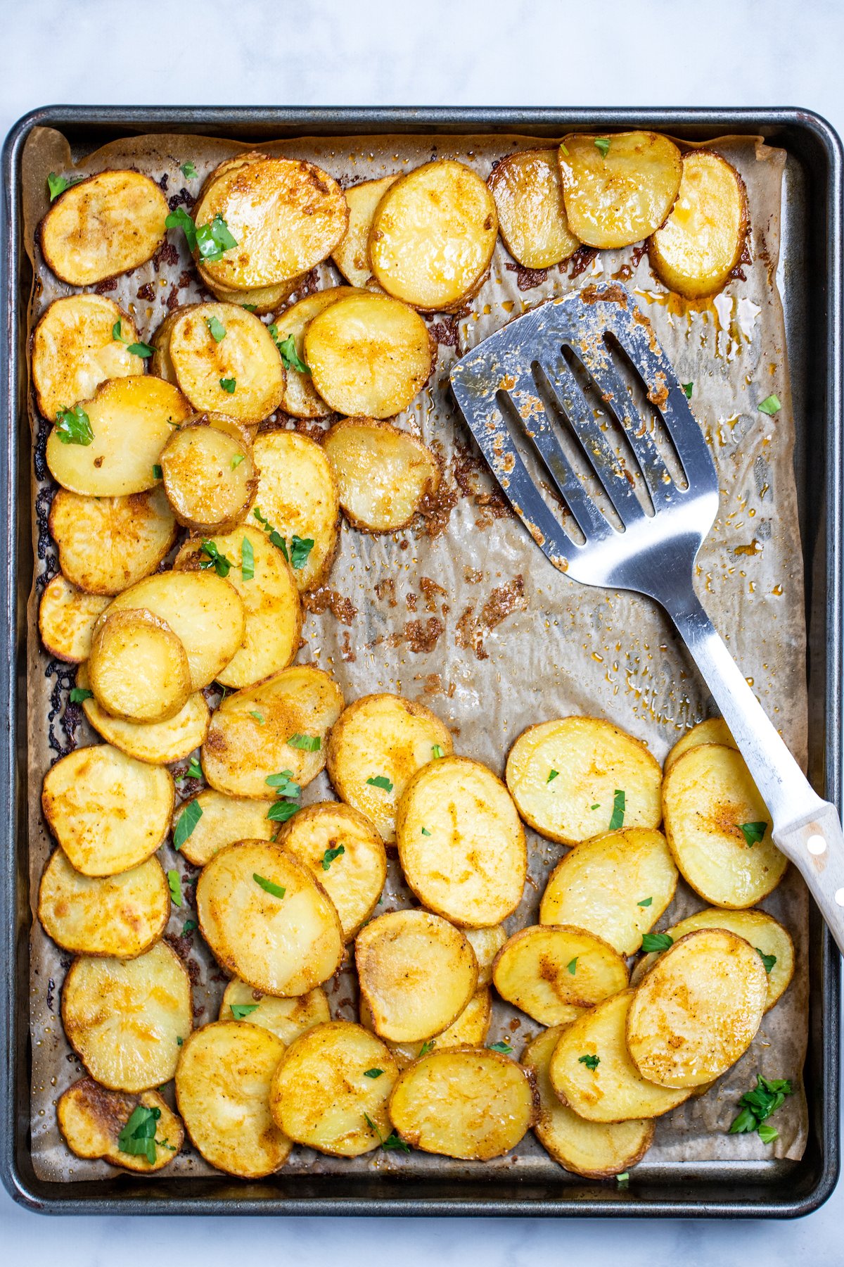 A sheet pan lined with parchment paper, with cooked baked potato slices topped with fresh parsley, and a spatula resting on the baking sheet.