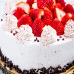 A strawberry chocolate cake on a wooden cake stand, decorated with mini chocolate chips and frosting dolloped on top with fresh whole strawberries.