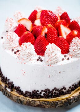 A strawberry chocolate cake on a wooden cake stand, decorated with mini chocolate chips and frosting dolloped on top with fresh whole strawberries.