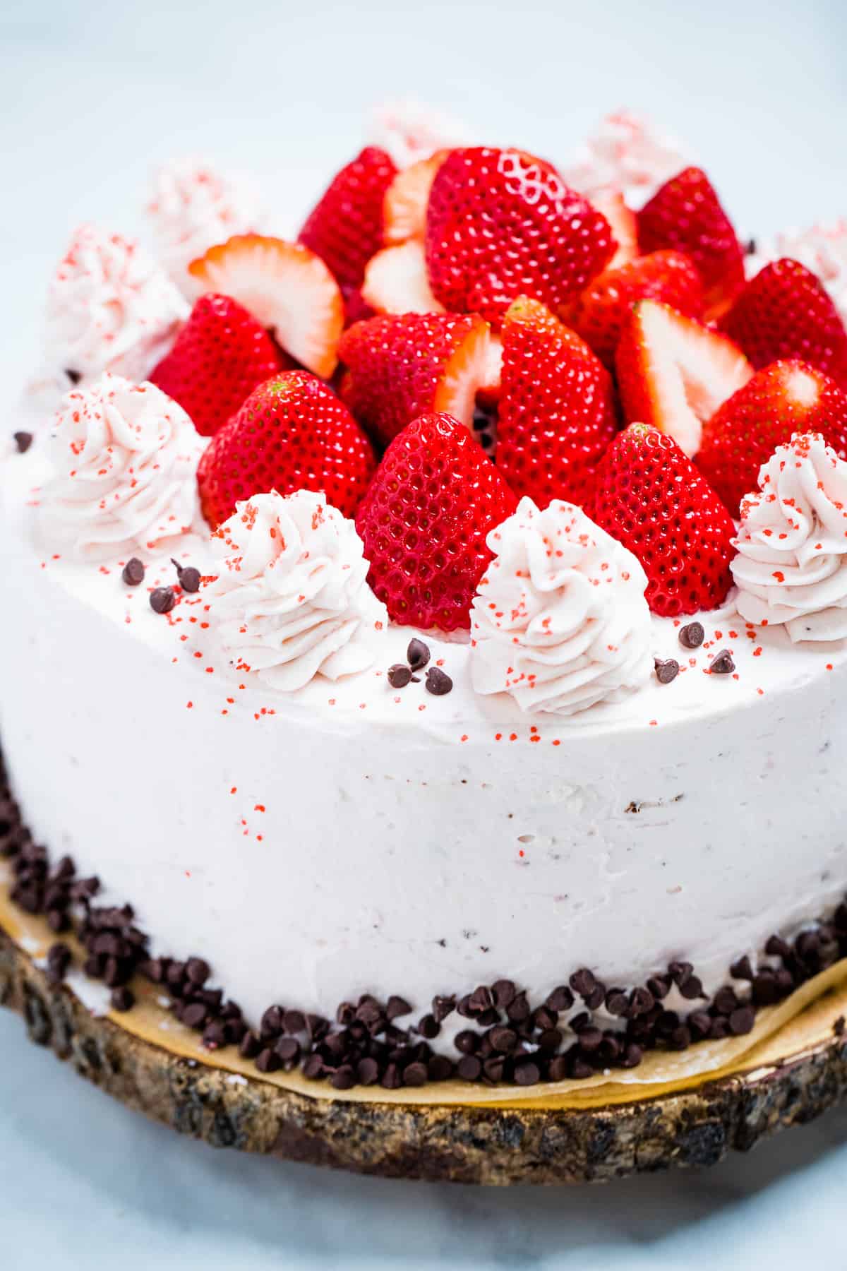 A strawberry chocolate cake on a wooden cake stand, decorated with mini chocolate chips and frosting dolloped on top with fresh whole strawberries.