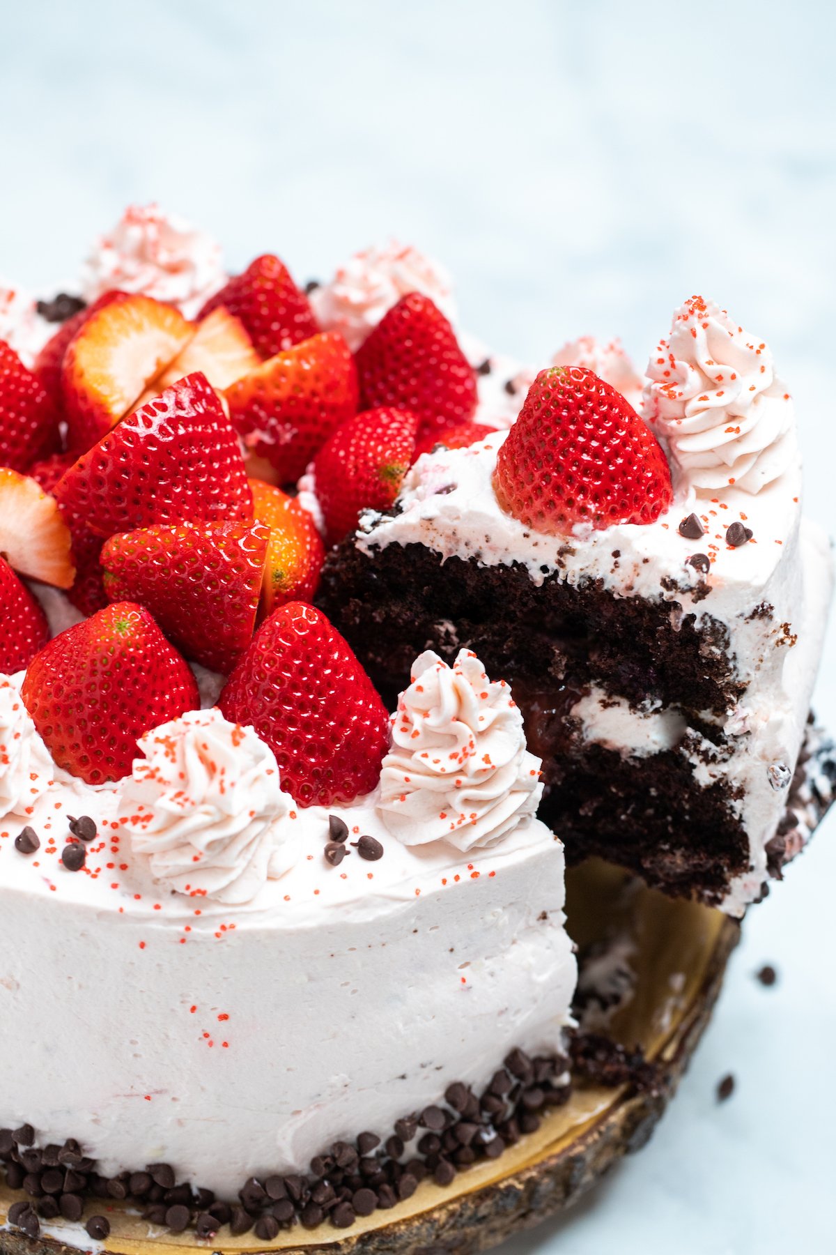 A strawberry chocolate cake on a wooden cake stand, decorated with mini chocolate chips and frosting dolloped on top with fresh whole strawberries, with a piece of cake being lifted up with strawberry filling on the inside.
