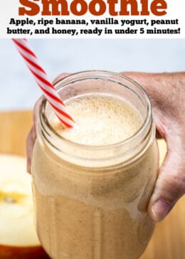 Pinterest pin with a hand holding an apple banana smoothie in a mason jar with a straw above a wooden cutting board with an apple cut in half behind it.