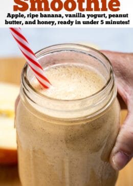 Pinterest pin with a hand holding an apple banana smoothie in a mason jar with a straw above a wooden cutting board with an apple cut in half behind it.