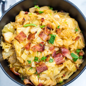 A bowl of crockpot cabbage topped with crispy bacon and parsley on a marble table.