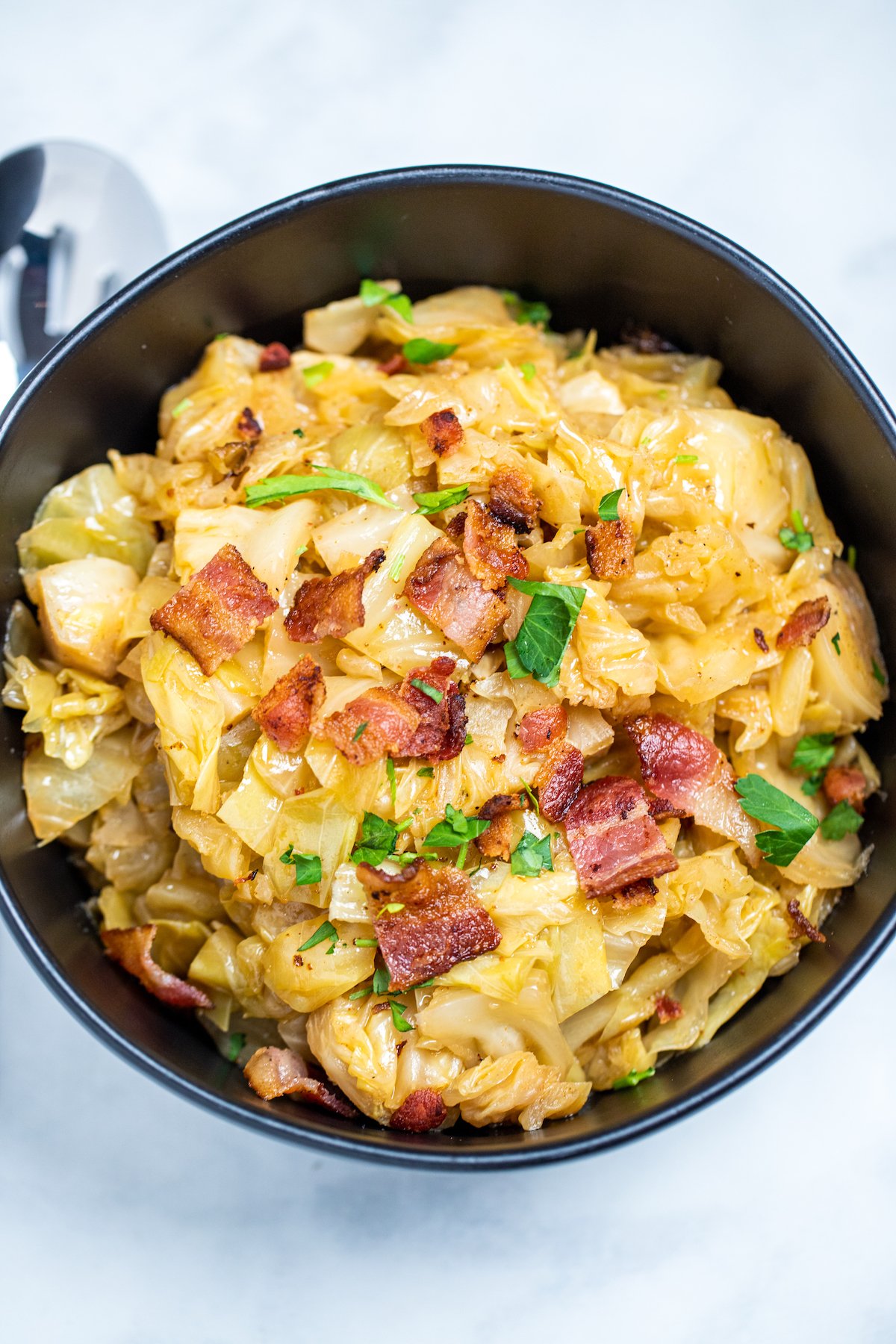 A bowl of crockpot cabbage topped with crispy bacon and parsley on a marble table.