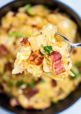 A closeup of a spoon holding a scoop of crockpot cabbage over a serving bowl, topped with crispy bacon and parsley.