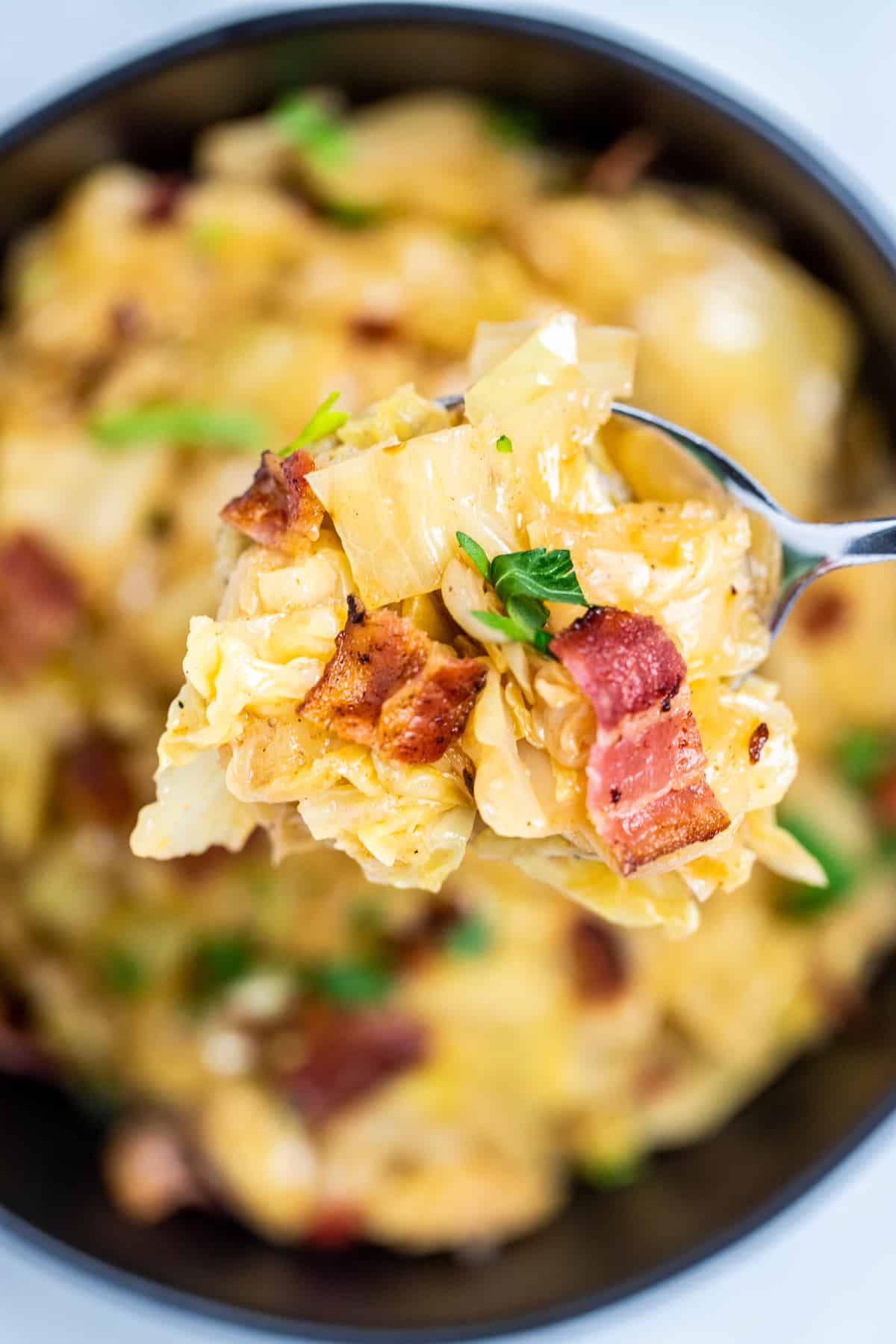 A closeup of a spoon holding a scoop of crockpot cabbage over a serving bowl, topped with crispy bacon and parsley.