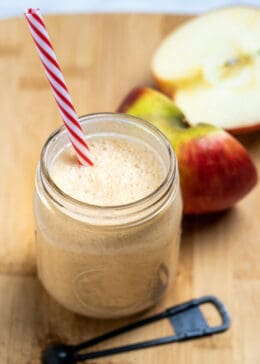 An apple smoothie in a mason jar with a straw sitting on a wooden cutting board with a teaspoon of cinnamon in front of it, and an apple cut in half behind it.