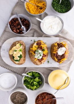 Three baked potatoes on a marble serving platter, with bowls of baked potato toppings next to it on the table.