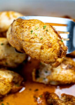 A seasoned fully cooked boneless skinless chicken thigh being lifted on a spatula over a baking dish of thighs.