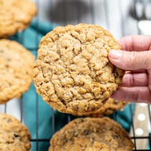 A hand holding a gluten free oatmeal cookie with cookies below on a cooling rack, above a kitchen towel.