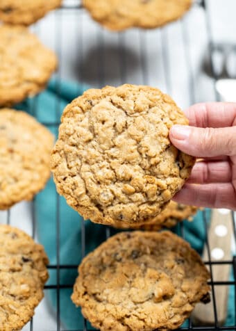 A hand holding a gluten free oatmeal cookie with cookies below on a cooling rack, above a kitchen towel.