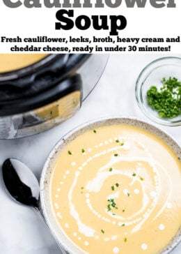 A bowl of cauliflower soup on a table in front of an instant pot, with cream drizzled on top and fresh chives next to a spoon.