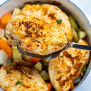 Chicken breast over potatoes and carrots in a dutch oven, with a spatula lifting one up.