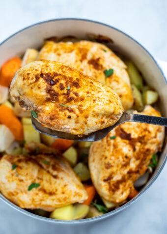 Chicken breast over potatoes and carrots in a dutch oven, with a spatula lifting one up.