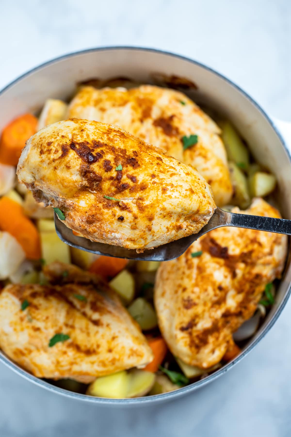 Chicken breast over potatoes and carrots in a dutch oven, with a spatula lifting one up.