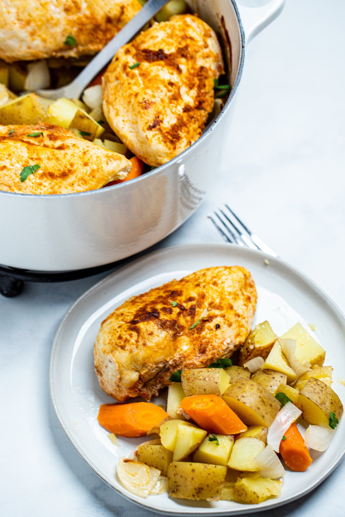 Chicken breast on a plate with potatoes and carrots and onions, in front of a dutch oven.