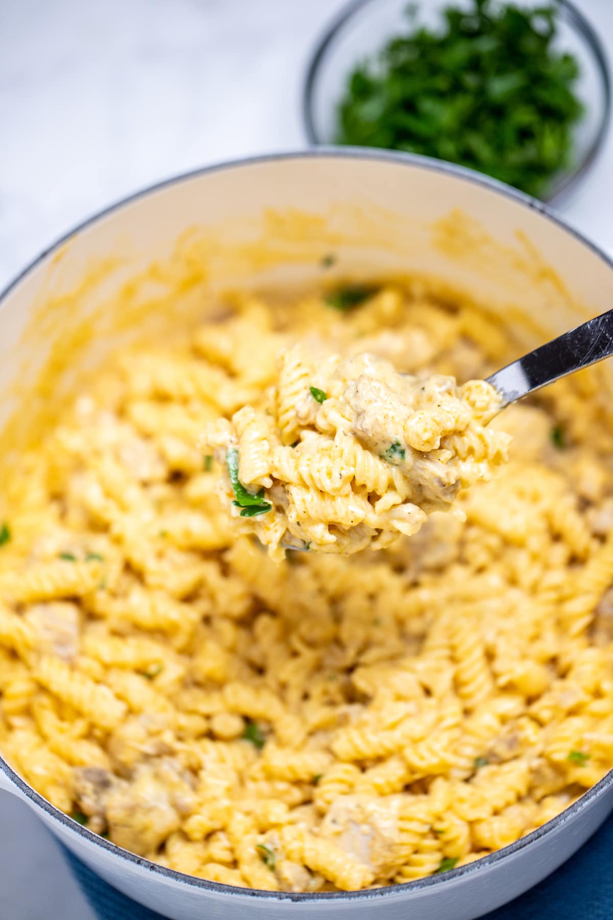 A spoonful of garlic parmesan chicken pasta being held over a dutch oven full of pasta, with a small bowl of chopped parsley in the background.