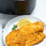 A piece of breaded tilapia on a plate with a lemon wedge in front of an air fryer basket.