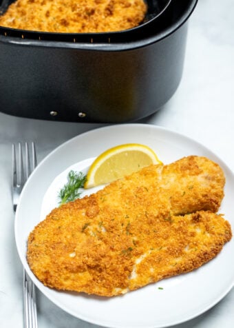 A piece of breaded tilapia on a plate with a lemon wedge in front of an air fryer basket.