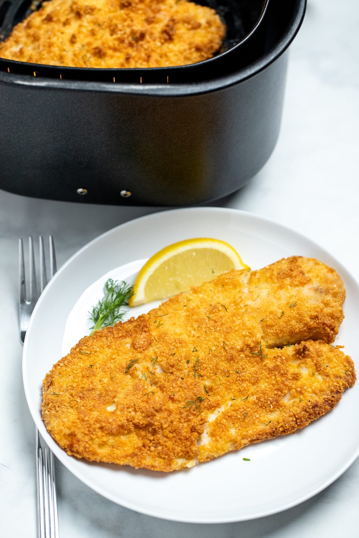 A piece of breaded tilapia on a plate with a lemon wedge in front of an air fryer basket.