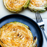 An air fryer cabbage steak on a plate next to a fork with more cabbage steaks on the table.