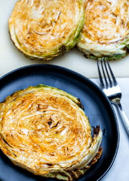 An air fryer cabbage steak on a plate next to a fork with more cabbage steaks on the table.